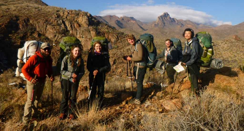 a group of students carrying backpacks pose for a photo in front of a desert landscape. there are mountains in the background
